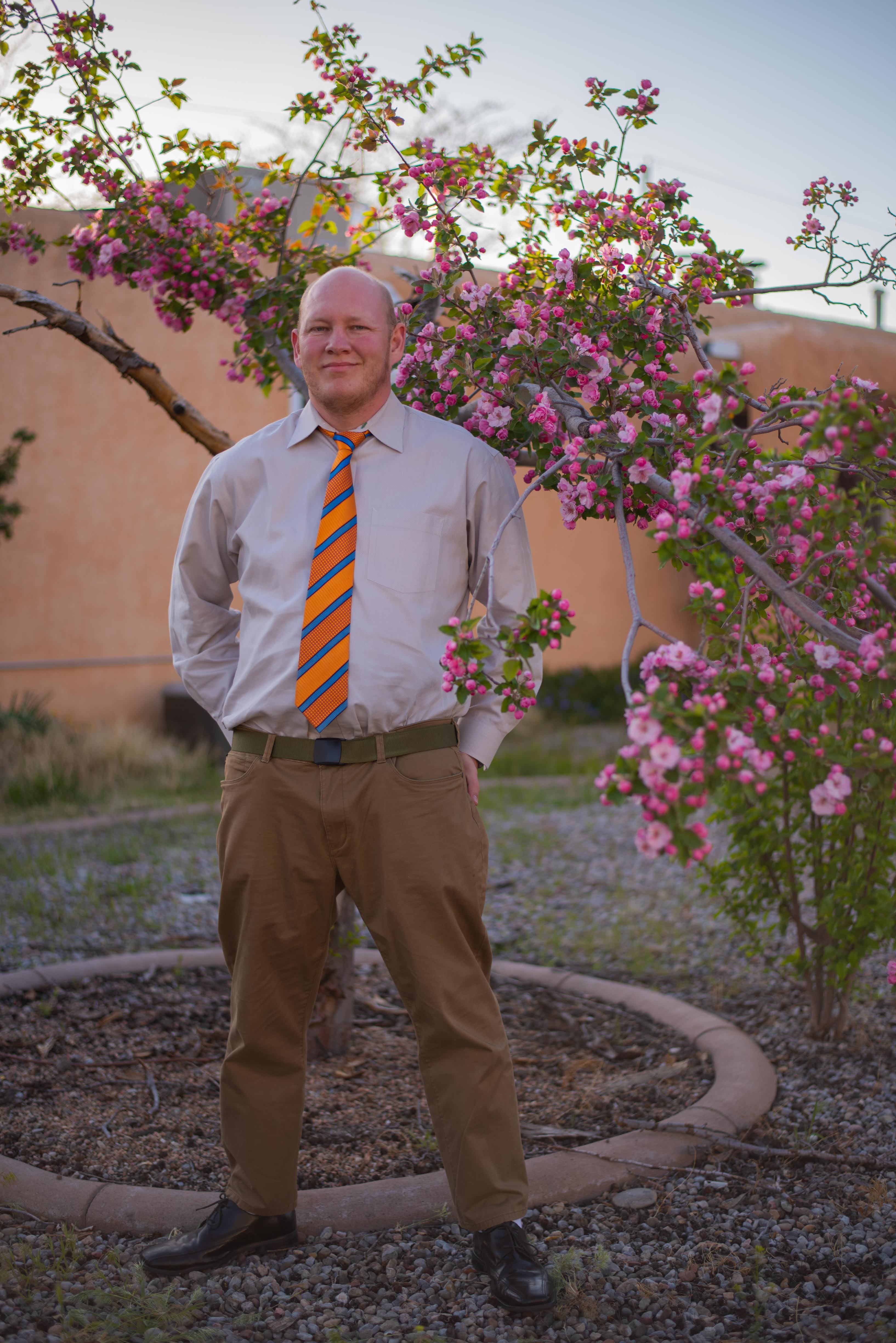 man in striped tie standing next to a apple tree in bloom with stucco house in background.